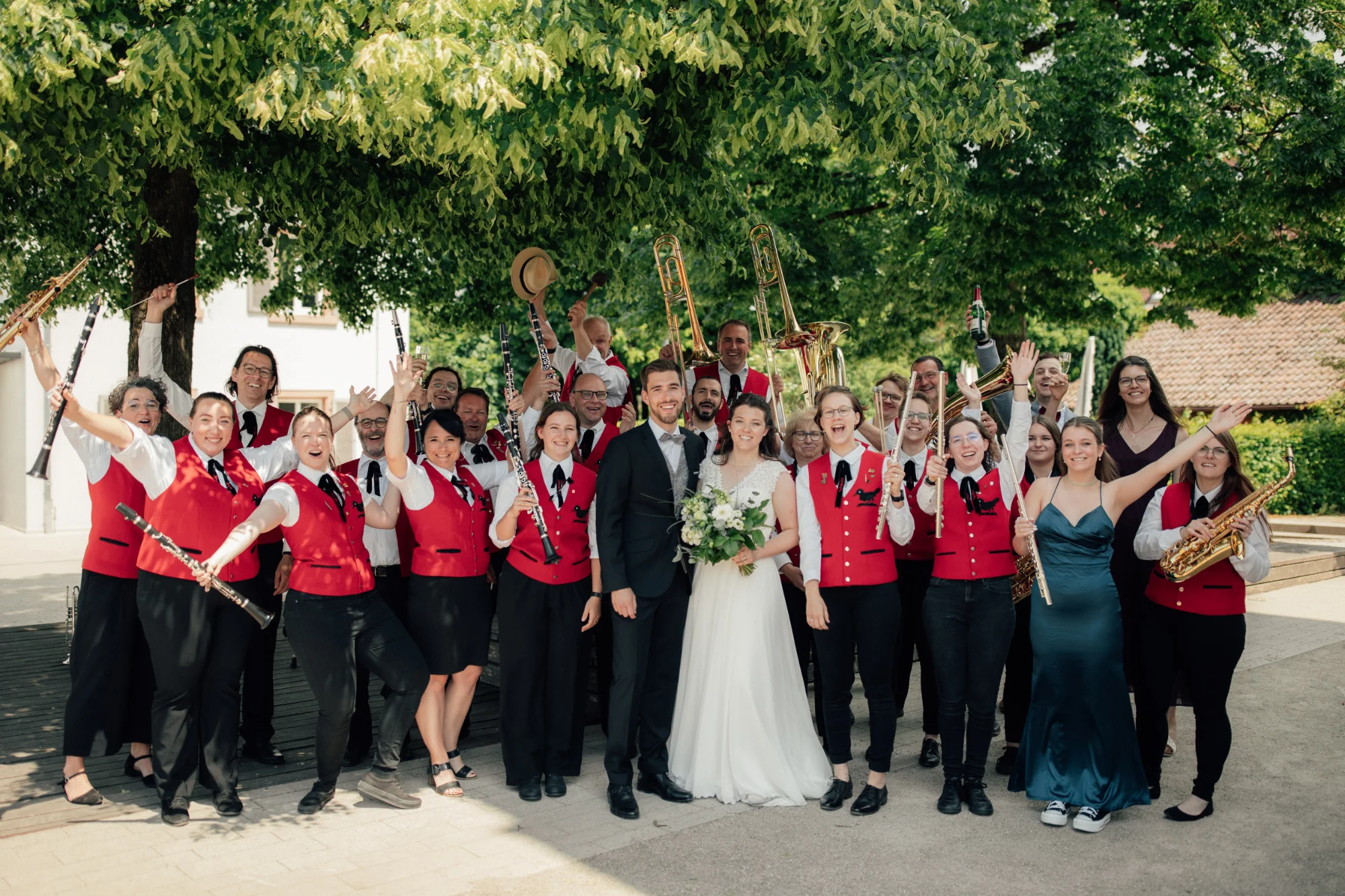 Musikerinnen und Musiker in roten Westen feiern mit ihren Instrumenten unter einem Baum mit dem Brautpaar