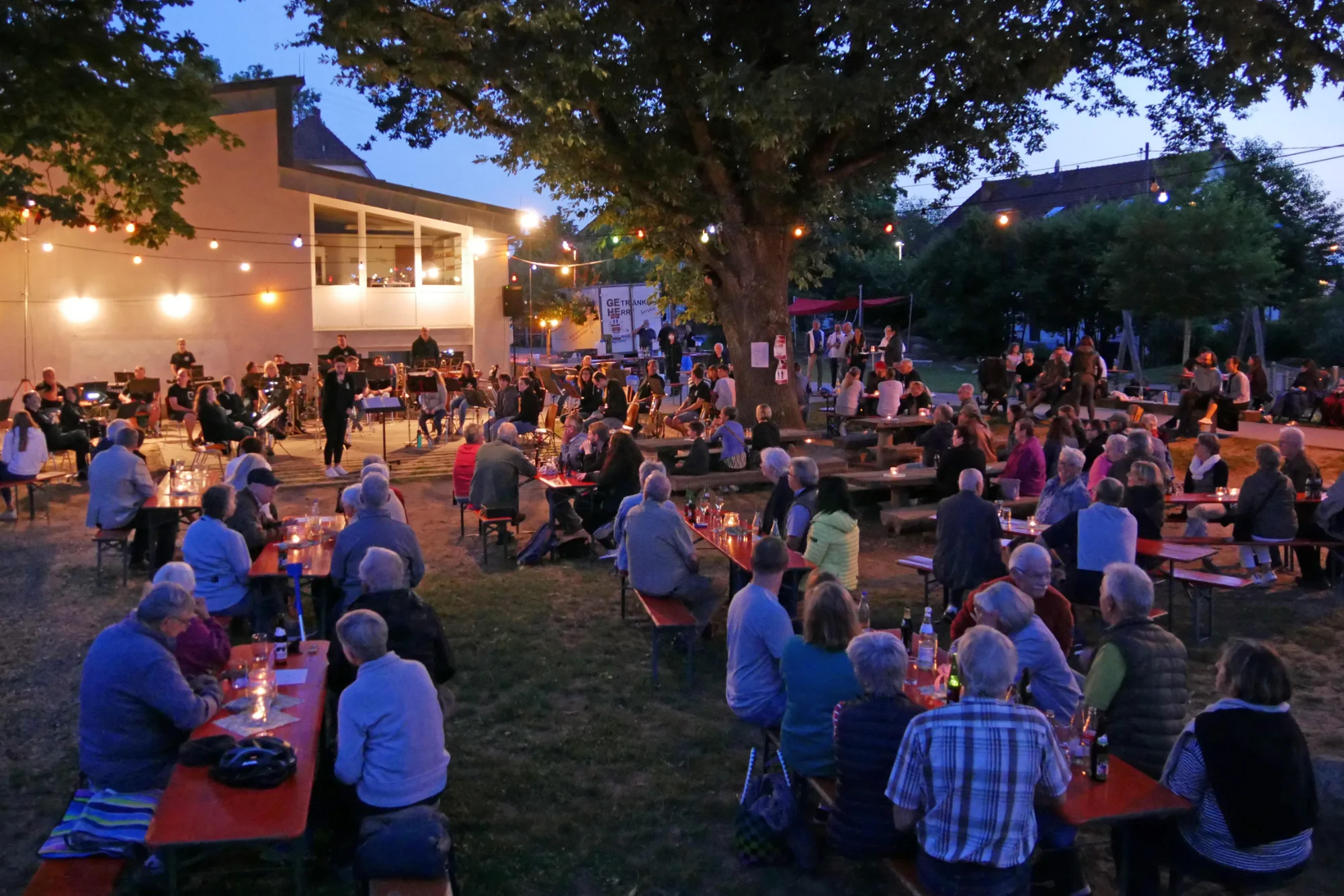 Gäste sitzen am Abend beim Open Air 2023 unter großen Bäumen und Lichterketten an Tischen