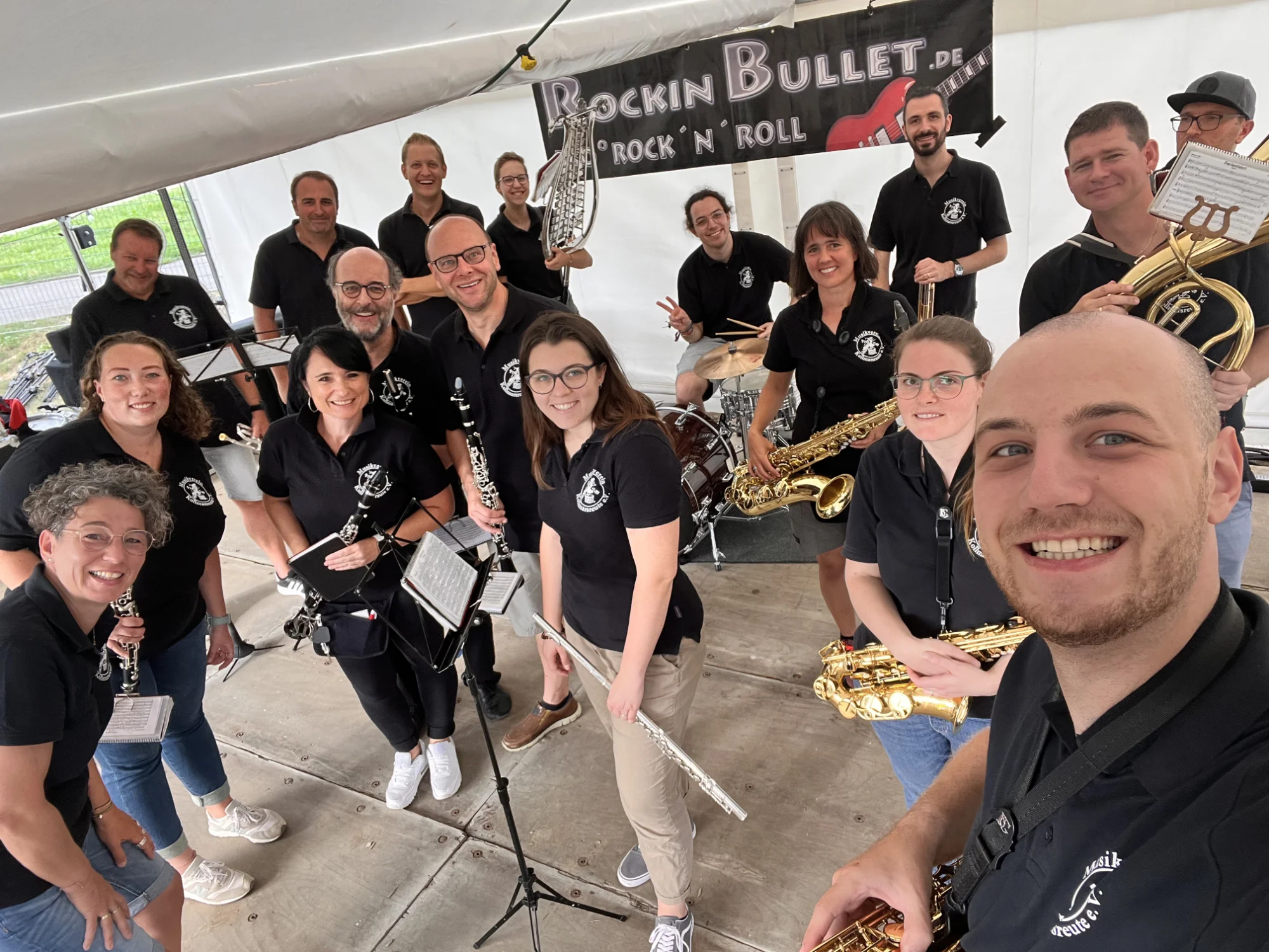 Musikerinnen und Musiker in schwarzen Vereinsshirts mit Saxophonen und Querflöten auf der Bühne, einer macht ein Selfie