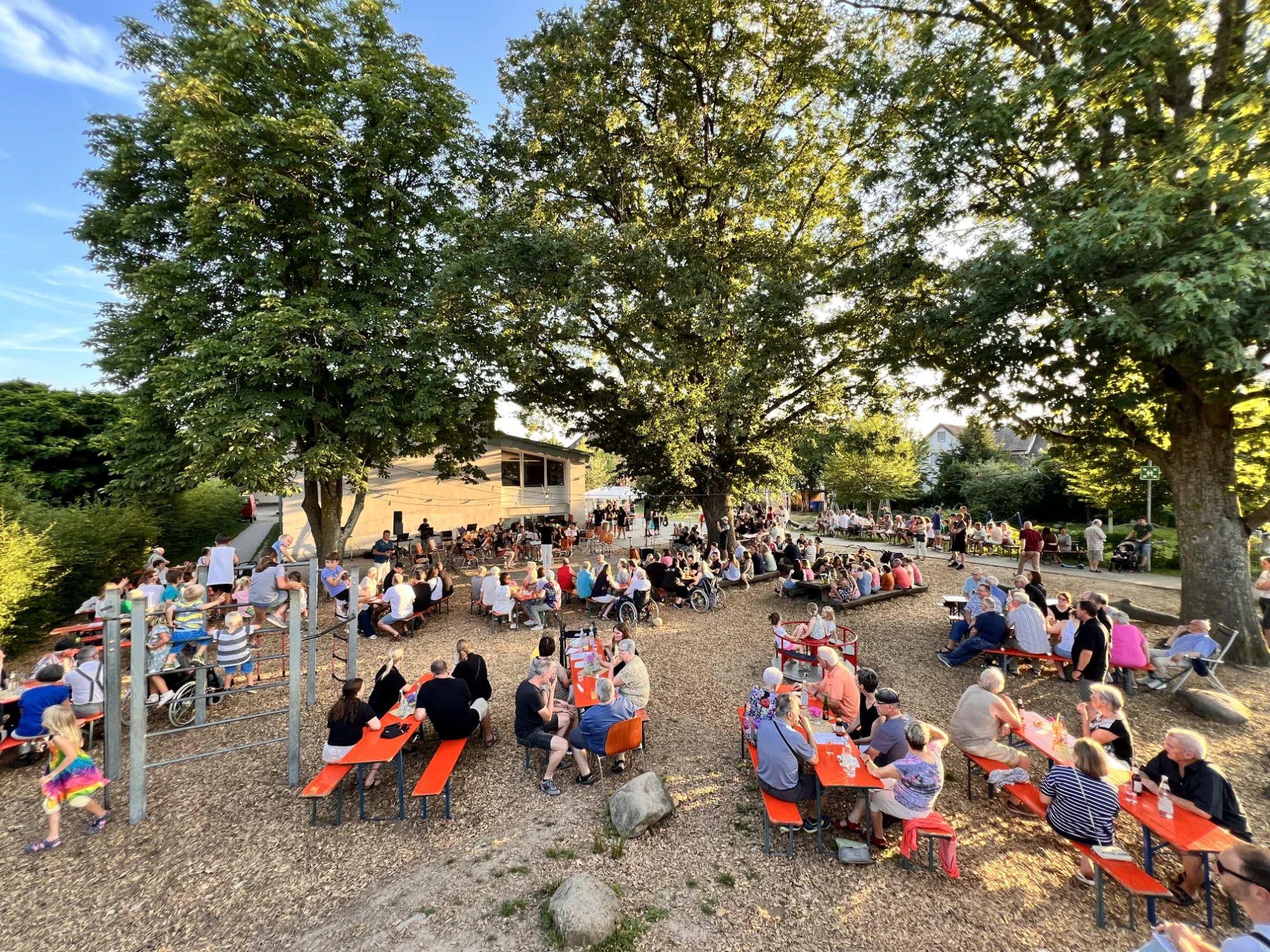 Zahlreiche Gäste sitzen beim Sommer Open-Air im Schulgarten an Bierbänken unter hohen Bäumen